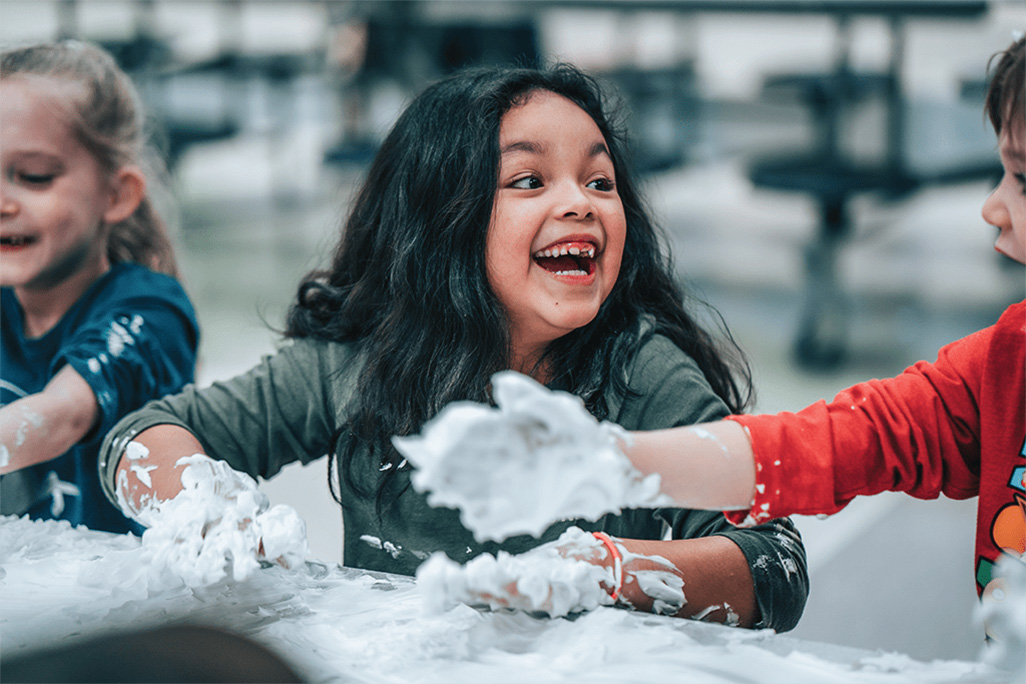 afterschool1-01-2 A girl sitting at a table looks to the person on her left, laughing as they play with shaving cream for a fun craft.