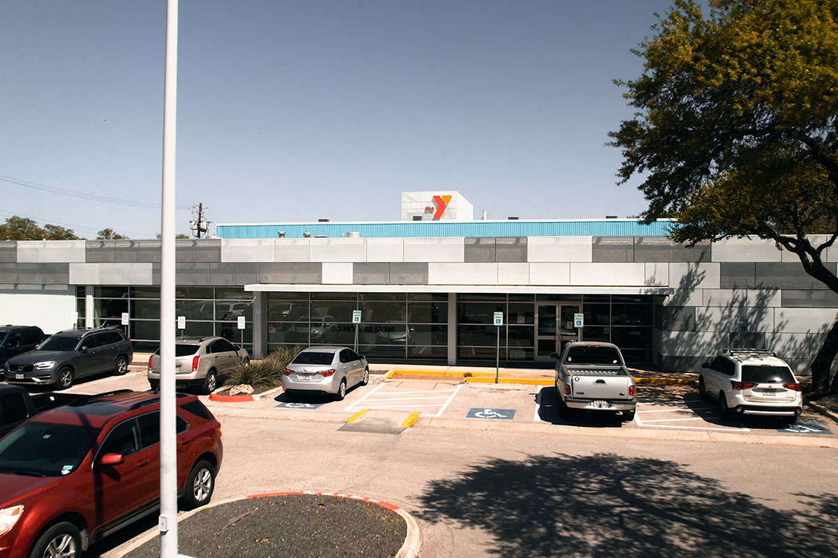 A two-story building with concrete facade. Windows line the front bottom story of the building with the entrance centered in the middle. Red and orange Y logo sits on top of building. Parking lot sits in front.