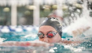 mastersswim-header An adult female member in a silver swim cap and goggles holds onto a kickboard while swimming laps at YMCA Camp Moody. Pool lanes and the natatorium’s windows are blurred in the background of the photograph.