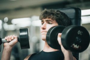 teenfitnessorientationform-header A teen male member wearing a black sleeveless shirt lifts dumbbells while seated, looking into the mirror in front of him. Weight room equipment is blurred in the background of the photograph.