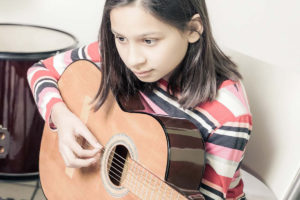 youthprograms-musicprogramsimage A girl in a striped pink shirt holding a guitar looks at the sheet of music in front of her as she is guided by her male adult music teacher in a blue shirt, also holding a guitar. A drumset and the walls of a music studio are blurred in the background of the photograph.