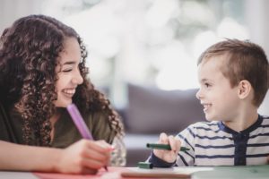 A teenage girl in a green shirt and a young boy in a striped blue shirt look at each other and share a laugh while coloring with paper and markers on a table. A window is blurred in the background of the photograph.