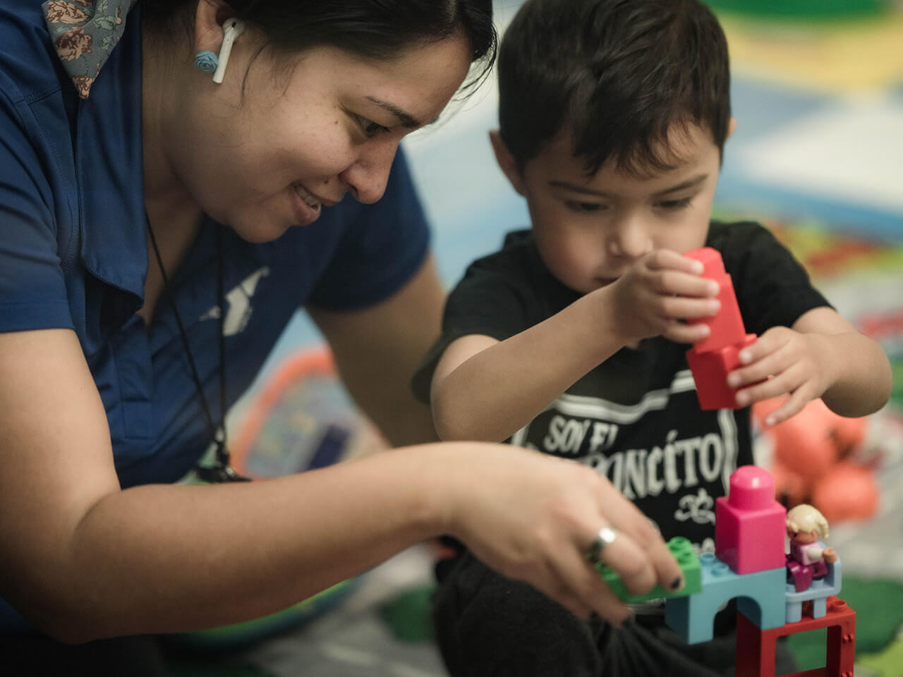 A woman and a young boy sit on the floor closely together. They are building with colorful building blocks.
