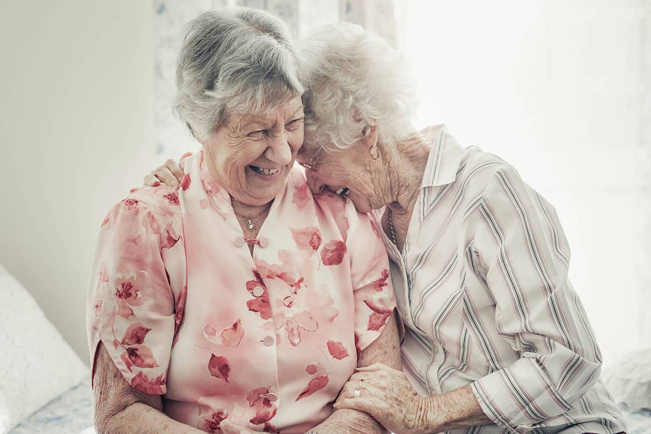 A senior woman in a pink flowered shirt and a senior woman in a striped white shirt laugh and embrace while seated together on a couch. The woman in the white shirt has her hands placed on the other woman’s shoulder and arm. A white wall and window are blurred in the background of the photograph.