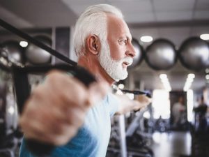 trxsuspensiontraining-overview A senior male member in a blue shirt leans forward into a position using a TRX suspension band. Weight machines and equipment are blurred out in the background of the photograph.