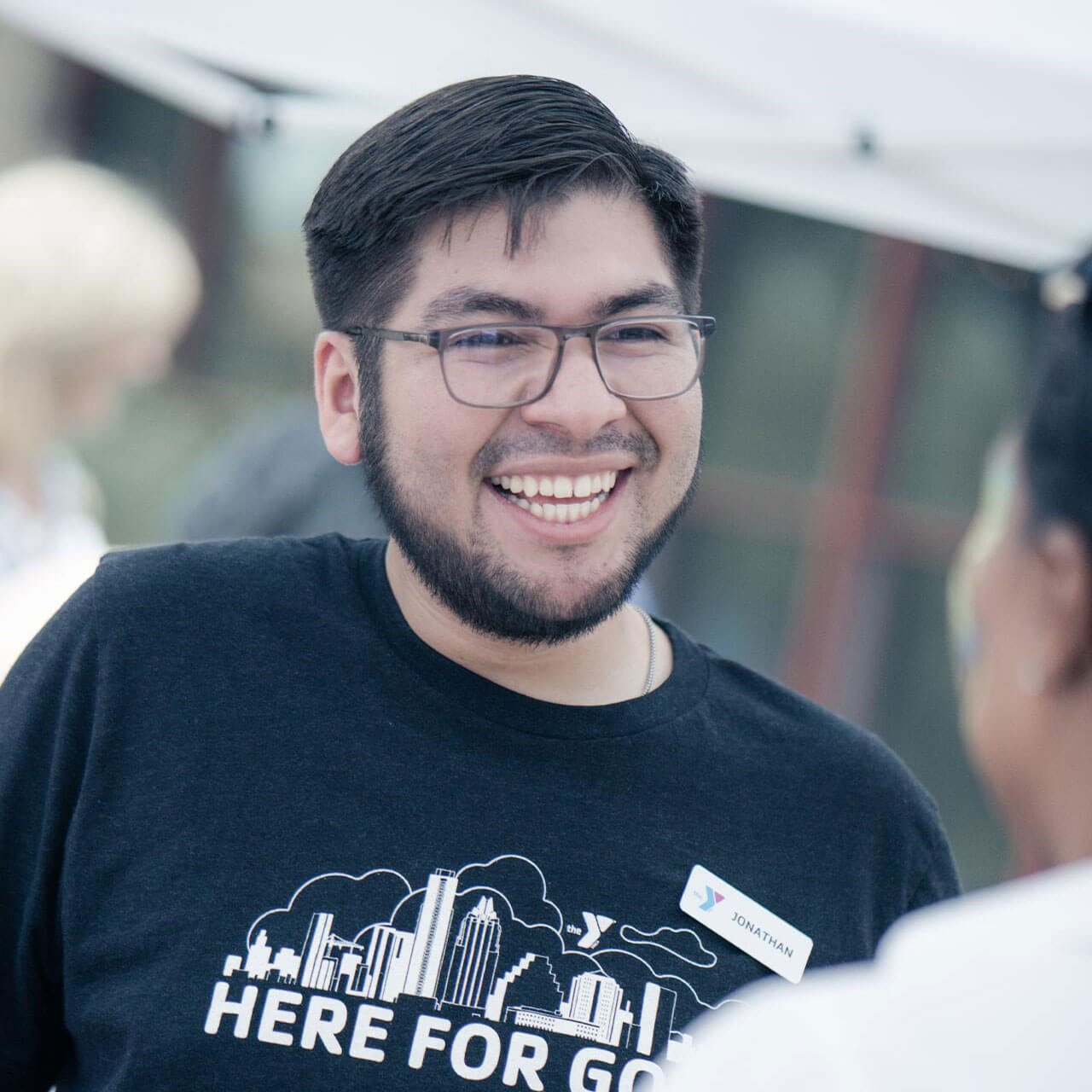 An adult male volunteer in a black shirt and glasses converses with a woman in a white shirt while standing together under a white canopy tent. An adult female and window covered building are blurred in the background of the photograph.