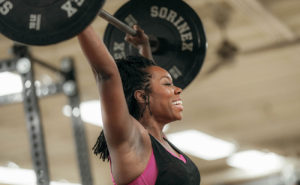 womensfitness-header An adult female member in a black sleeveless shirt and pink sports bra smiles as she lifts a bar of weights over her head during her power clean exercises. The ceiling of the weight room is blurred out in the background of the photograph.