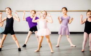 youthdance-header Six girls wearing different colored leotards and tutus look up to their dance instructor and while doing the chicken dance. A dance studio is blurred in the background of the photograph.