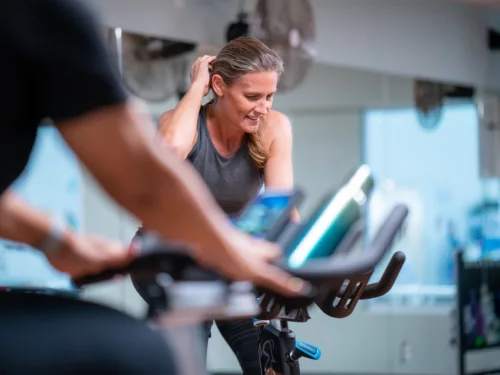 benefits-slider-group-exercise A woman smooths her hair back as she exerts herself on a stationary cycle in a group fitness class.