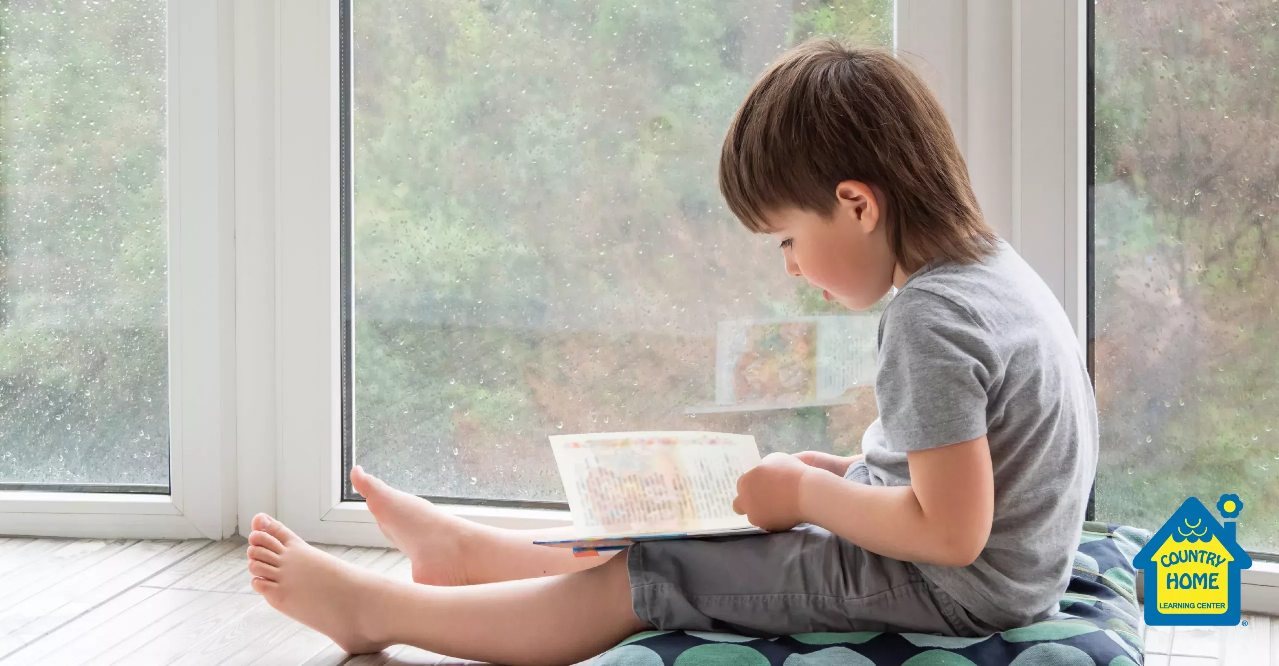 Child reading a book by the window on a rainy day.