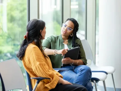 Two women sit near a window in close conversation with one another. One woman holds a clip board.