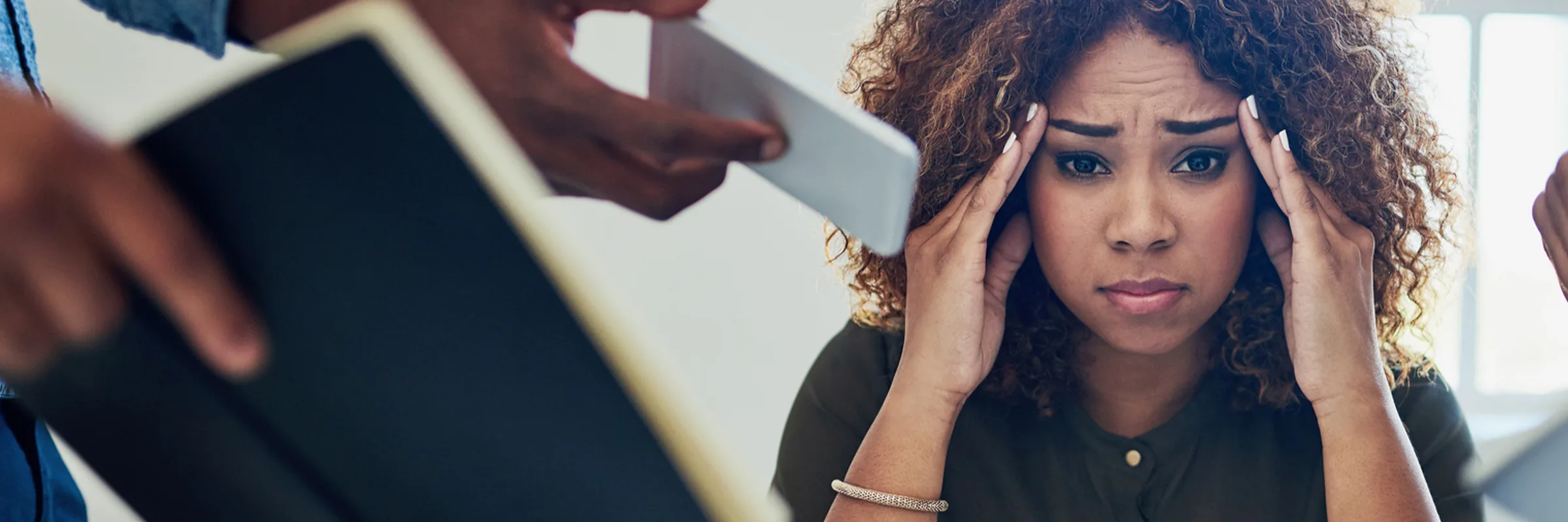 A woman sits at a desk with her hands on her temples. People surround her holding devices, papers, phones, and more, communicating that they all need her attention.