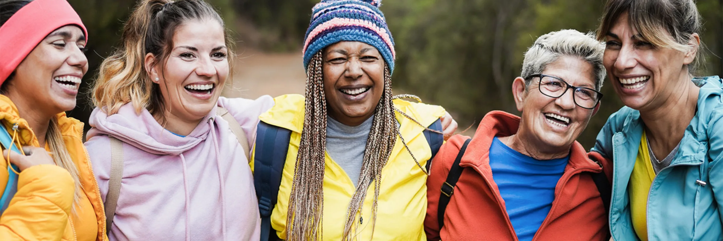 A group of friends stand smiling, arm-in-arm, outdoors on a hiking trail. They are wearing brightly colored clothing.