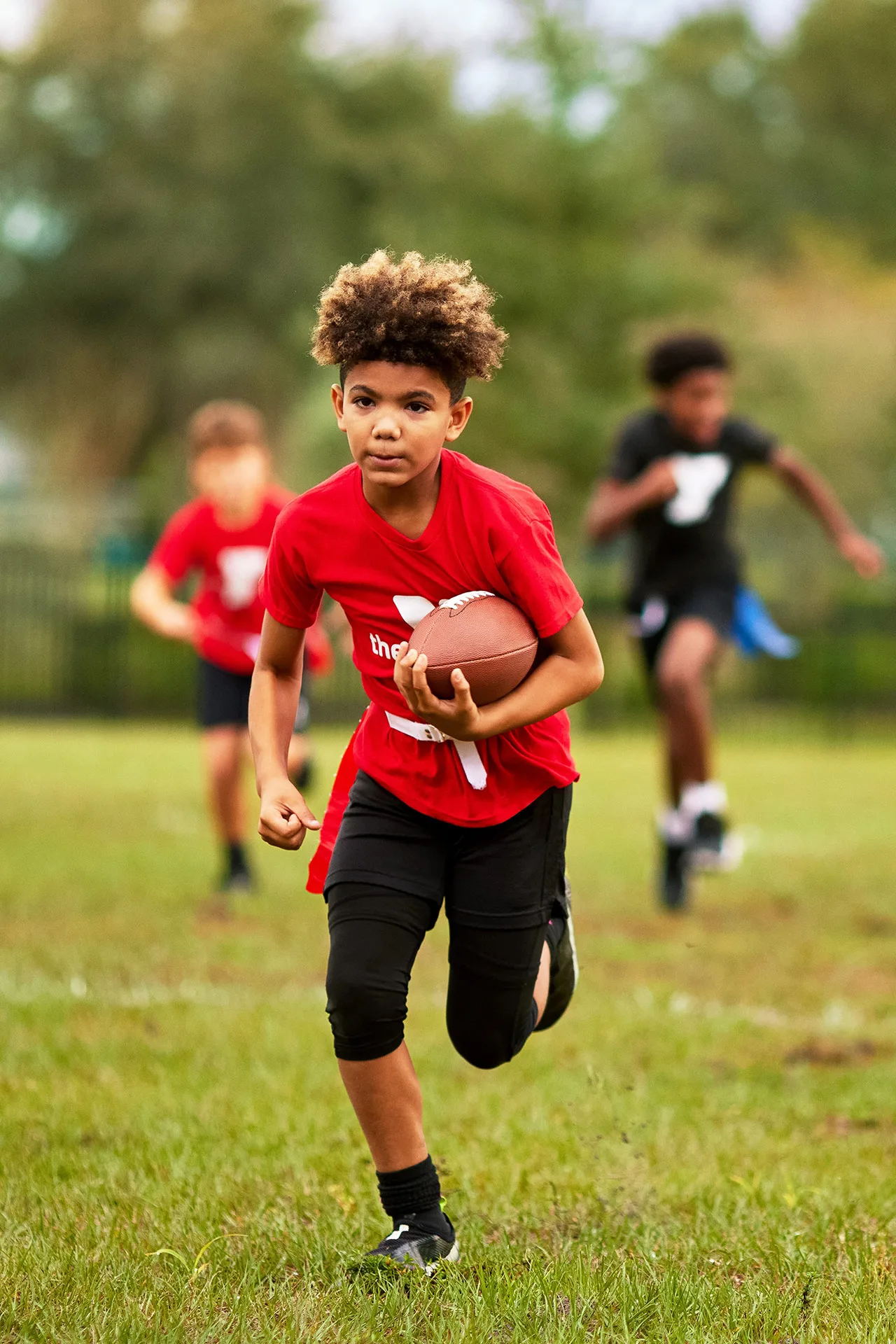flag-football-overview-vertical A child runs across a field holding a football. Children chase him in the background.