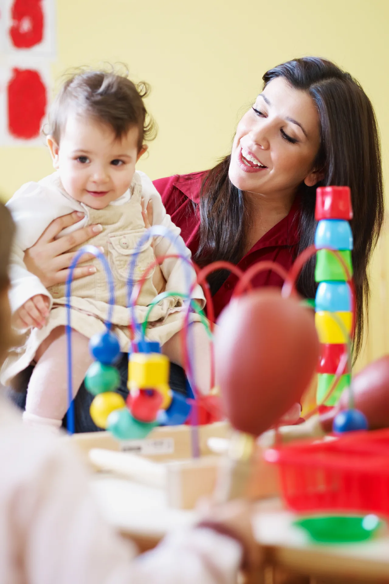 a woman sits at a table piled with colorful toys and holds a baby on her lap