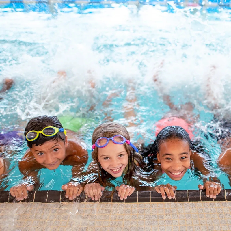 Children wearing goggles and colorful swimsuits cling to the edge of a pool and practice their kicking. The kids are smiling and having fun.
