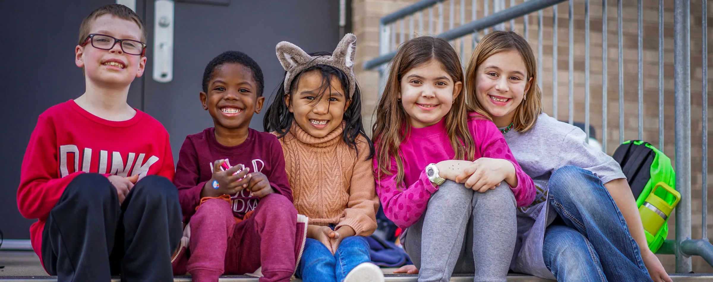 Blog-Banner-Oak-Hill-2400×800 Five children sit together on the steps of their school smiling at the camera.
