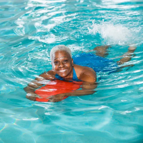 adult-swim-lesson-square-2 A woman smiles in a pool as she swims using a flotation device.