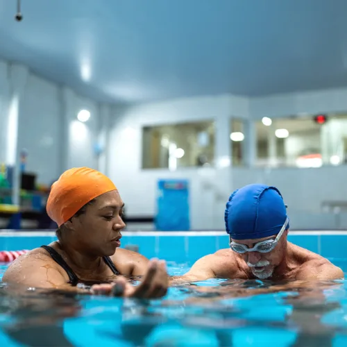 An adult man bends his head toward the water as a woman beside him coaches him in a swimming pose.