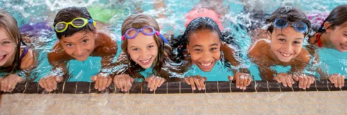 swimming-lessons-aquatics-header Children wearing colorful swimsuits and goggles smile at the edge of a pool.