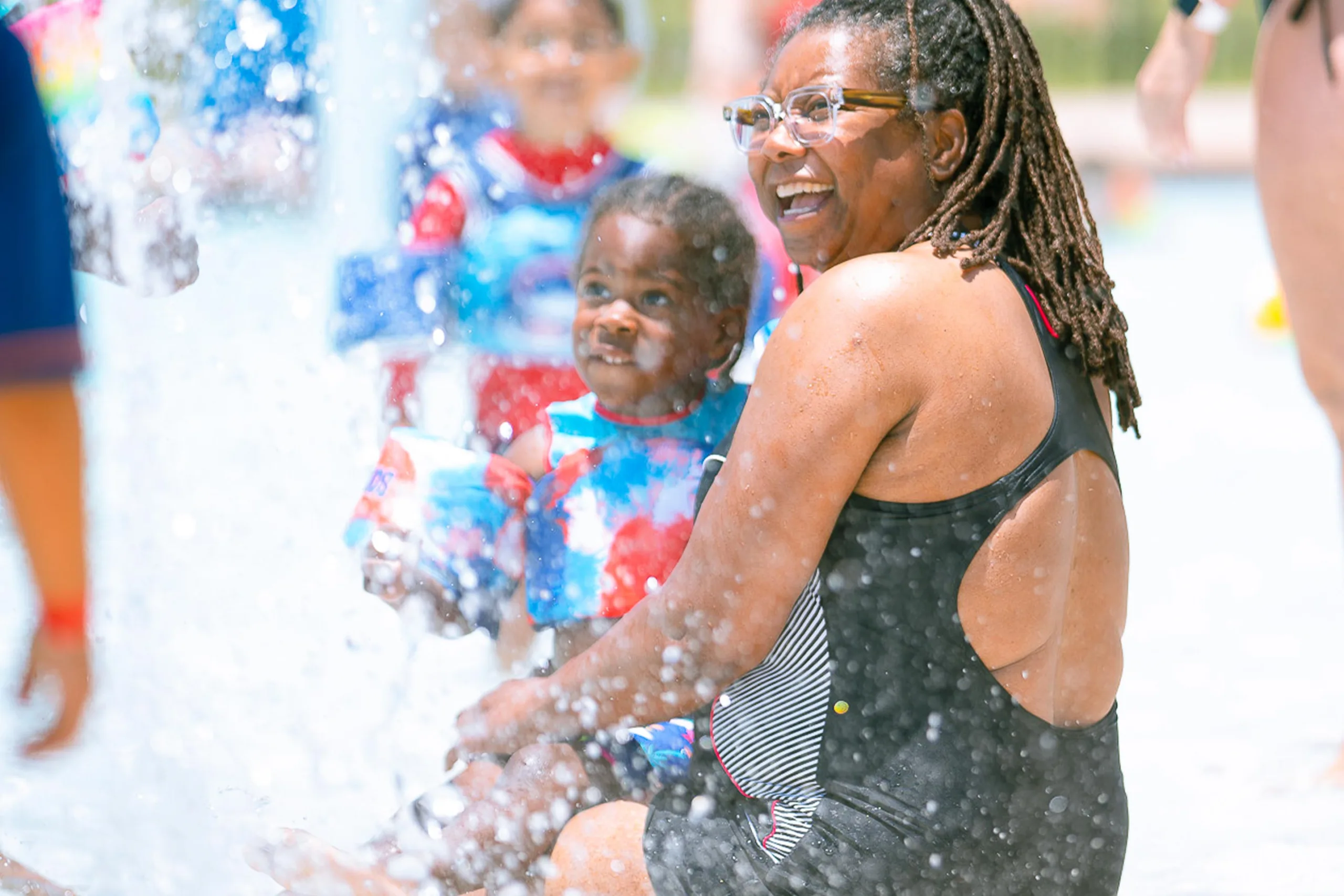 swimming-lessons-sub-header A parent and child smile and splash in a pool on a sunny day.