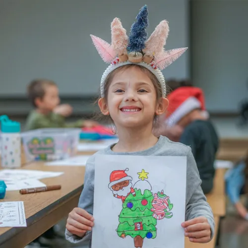 winter-camp-art-square A smiling girl with a winter tree headband holds up her holiday art