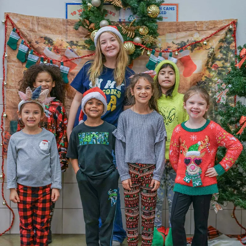 A group of children smile at the camera next to a Christmas tree in holiday themed outfits