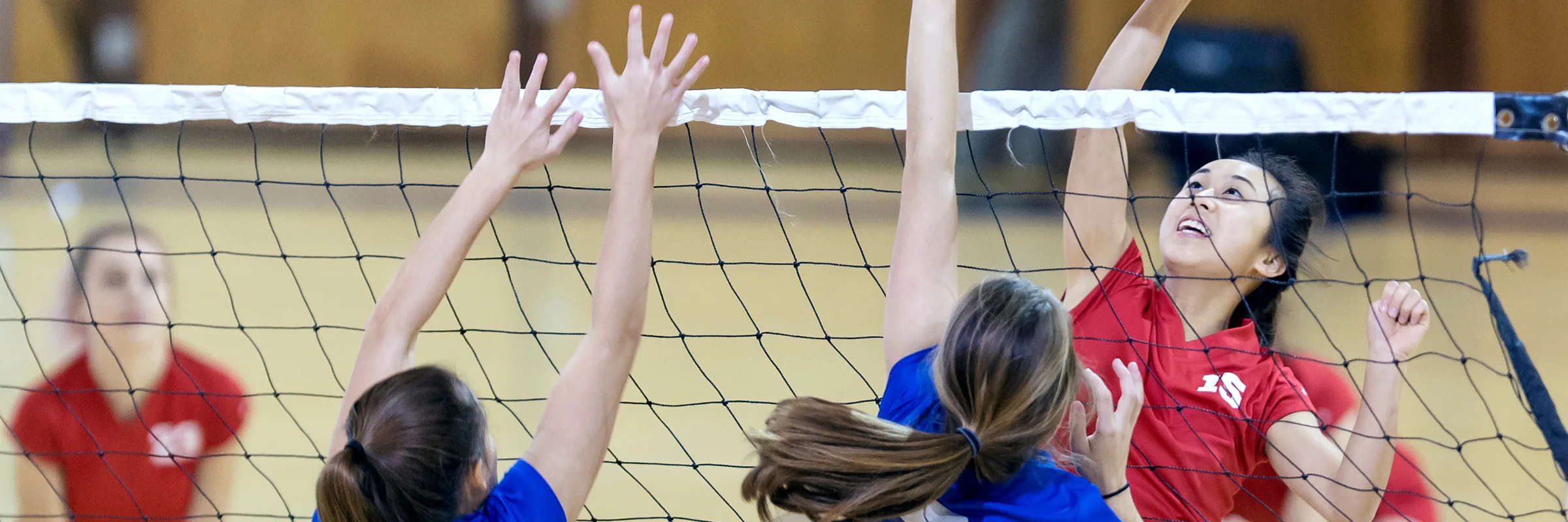 teen girls jump in the air at the net during a volleyball game.
