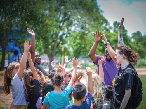 Group of children and counselors raising their hands together during an outdoor summer camp activity in a park.