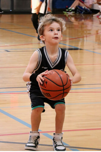 Young child focuses on making a basketball shot during a YMCA youth basketball game, learning skills and sportsmanship.