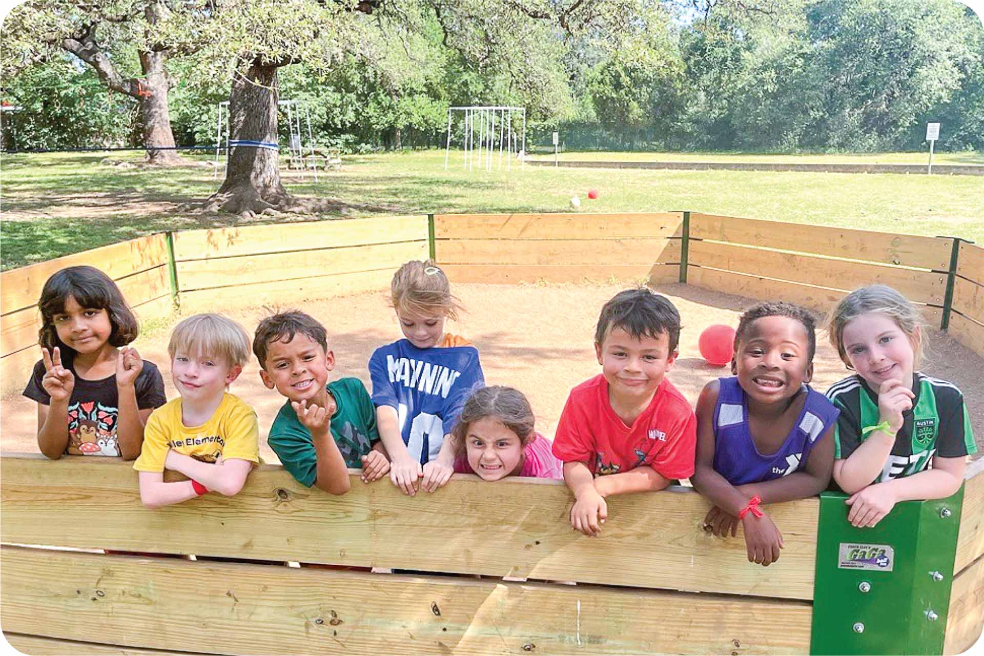 A group of eight diverse children stand together in a gaga pit outside with a ball behind them
