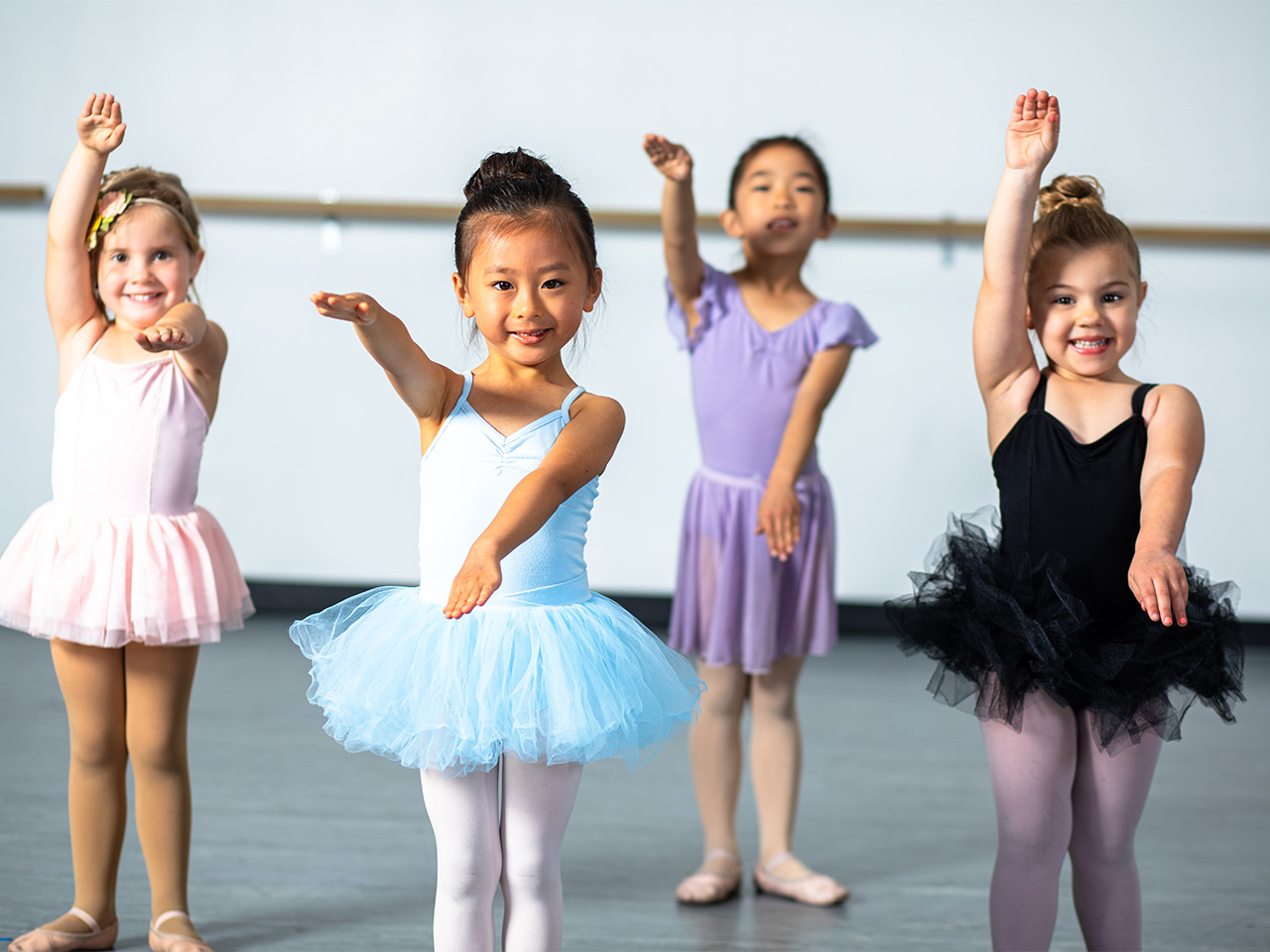 youth-dance-classes-ballet-lessons-horizontal Small children smile in tutus while practicing in a ballet dance class.