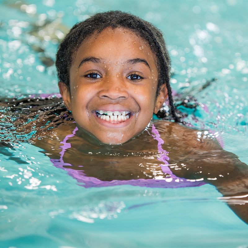 A child smiles in a pool during swimming classes to learn stroke introduction.
