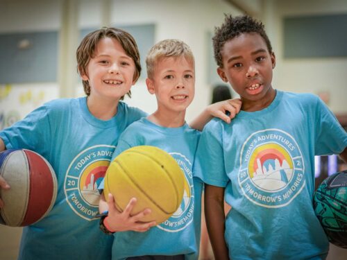 Three children smiling and holding basketballs during an indoor summer camp sports activity.