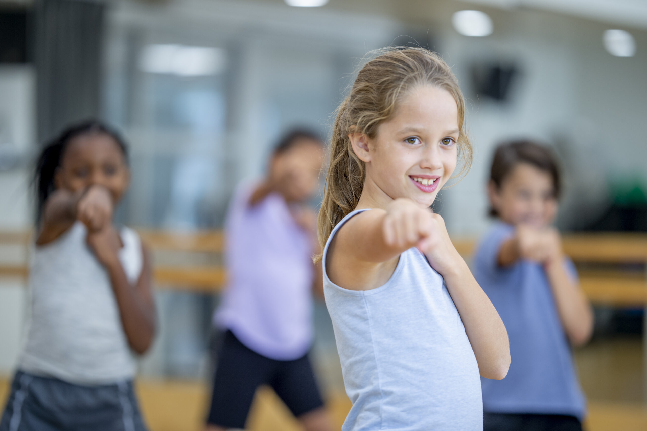A small group of school aged children stand in a gymnasium with their feet apart, one arm tucked in to their chest and the other outstretched in a fist as they participate in a boxing lesson. They are each dressed comfortably in athletic wear as they focus on increasing their strength.
