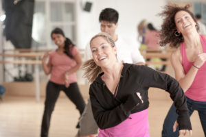 A multi-ethnic group of men and women in a dance fitness class in an indoor gym studio, wearing exercise clothing, and moving to the music.