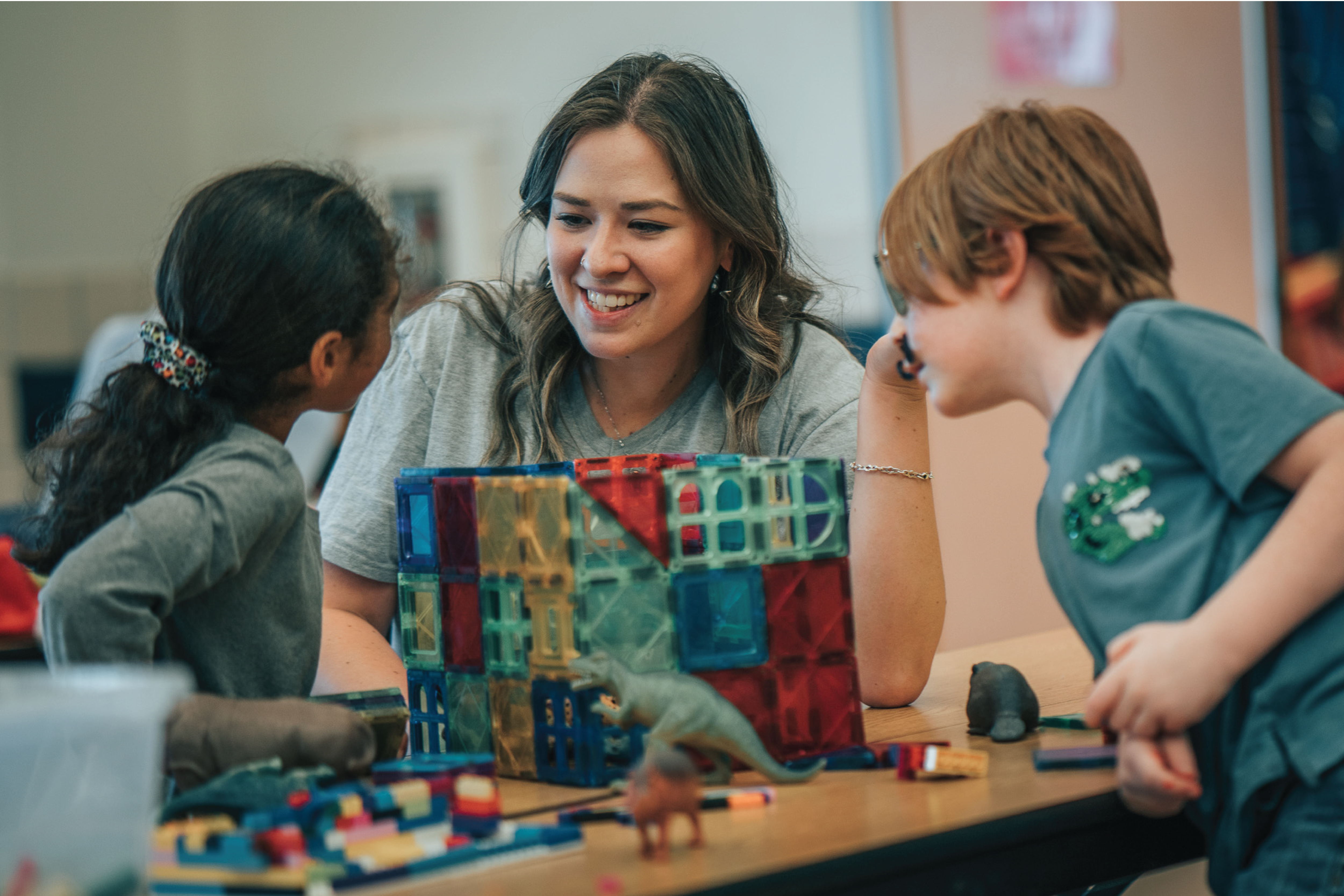 A youth development leader sits at a table with two children playing with colorful magnet tiles. The teacher is looking and smiling at one of the children while the other child looks on from the other side of the table.