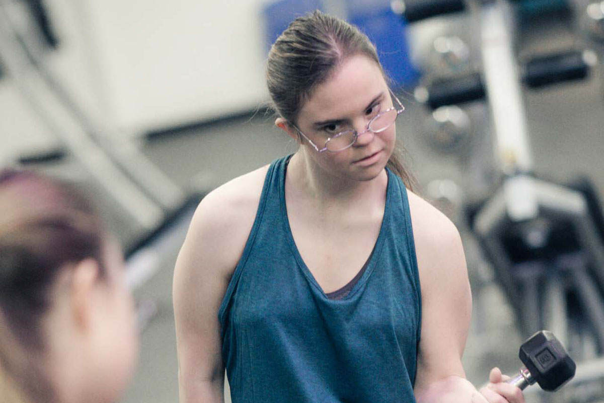 A female member in green sleeveless shirt and black pants and glasses looks into the mirror with focus as she does dumbbell curls. Weight machines are blurred in the background of the photograph