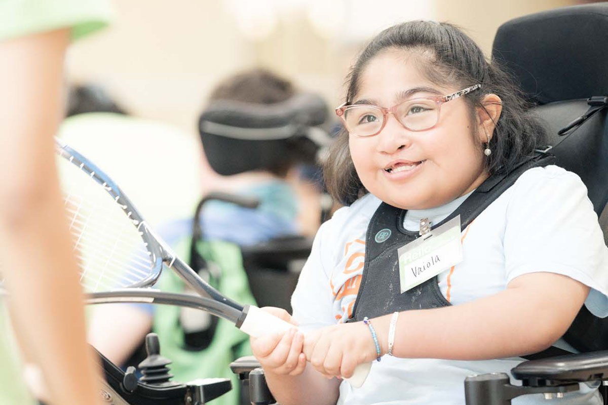 A young girl with glasses sits in a wheelchair and smiles while holding a tennis racket