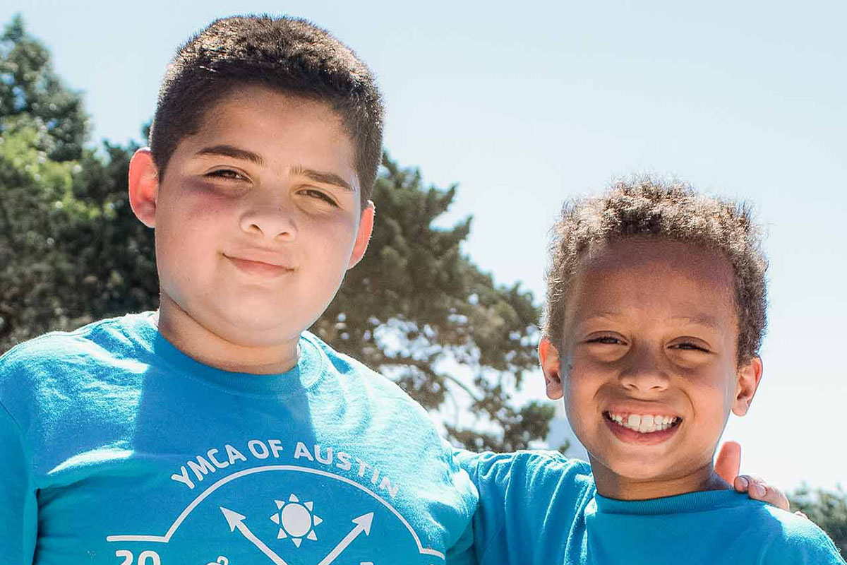 Two young, male campers in blue shirts put their arms around each other and smile for the camera while standing at a sand volleyball court in Zilker Park. Trees and the Austin skyline are in the background of the photograph.