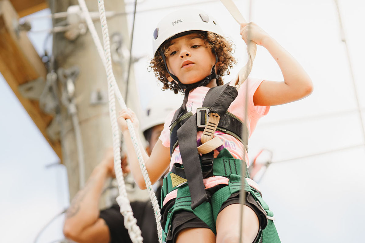 A young girl in a pink shirt and white helmet walks across the ropes course at YMCA Camp Moody, looking down below her. The ropes course and cloudy sky are blurred in the background of the photograph. 