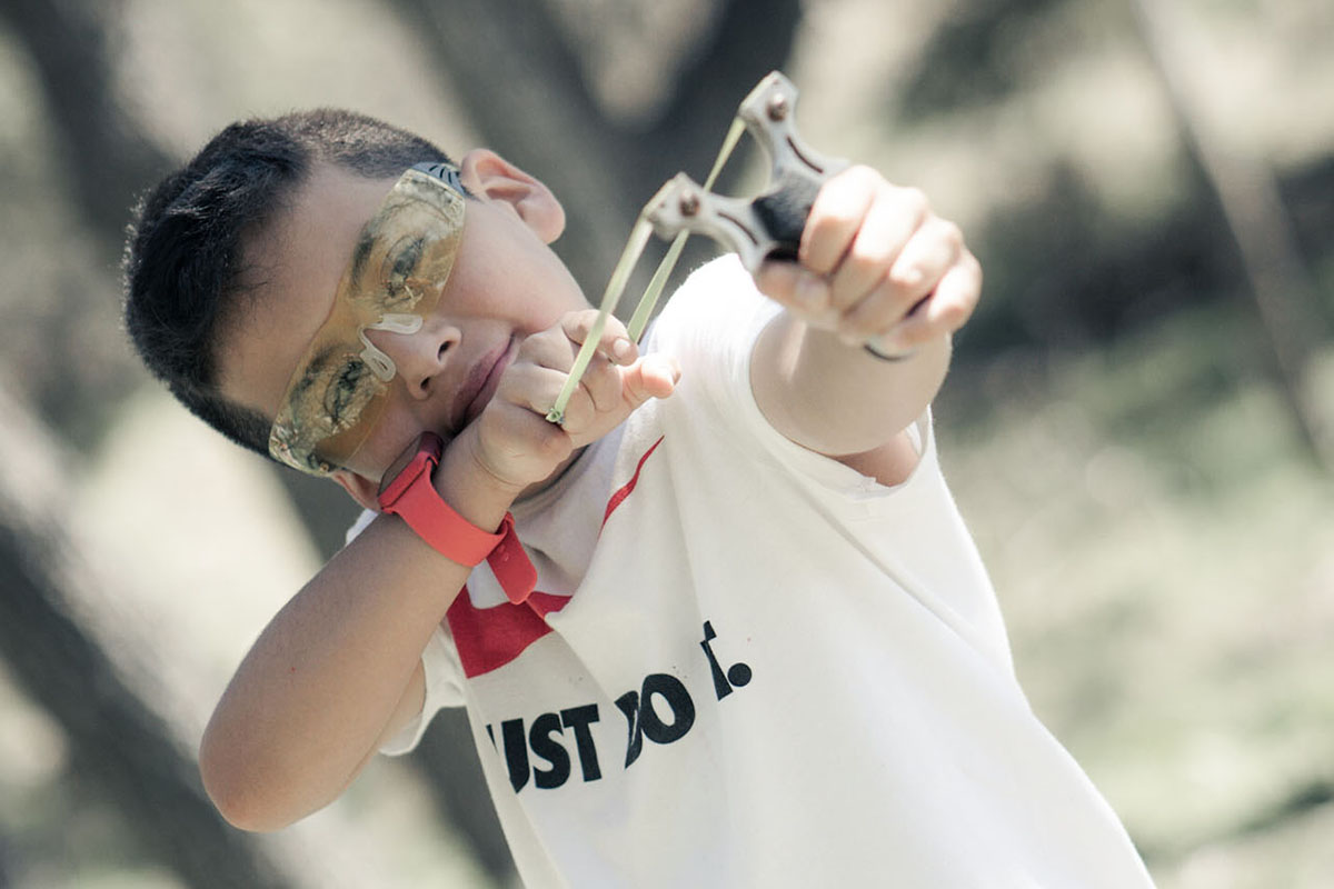 A young boy in a white shirt wearing protective eye gear pulls back on a slingshot