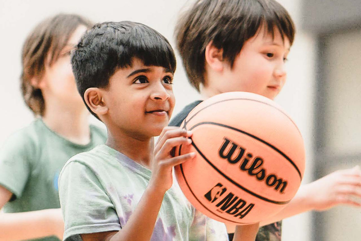 A young boy in a rainbow tie dye shirt dribbles a basketball and listens to instructions from his camp counselor. Other young, male campers in black and green shirts and the walls of the basketball gym are blurred in the background of the photograph.