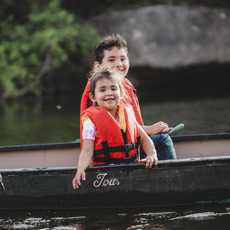 Two children sit in a canoe in water. They are wearing bright orange life vests. One of the children is reaching toward the water. Both children are smiling and looking at the camera.
