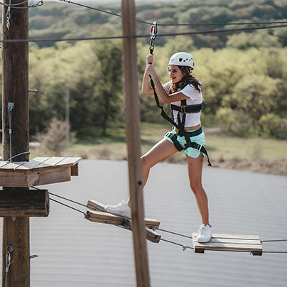 A teenage girl wearing a helmet and safety harness navigates a wooden high ropes course. She is smiling as she crosses the course. Trees are distant in the background below her.