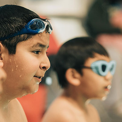 Children sit at the edge of a pool looking at their instructor who is not pictured. One boy has his goggles on his forehead. Another boy is wearing his goggles over his eyes.