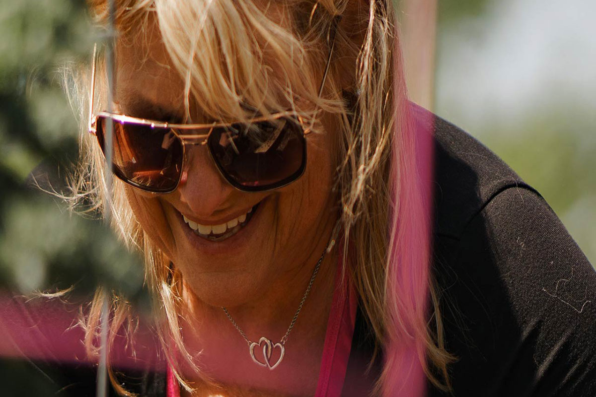 A female member in a black long sleeved shirt and sunglasses smiles as she looks down at a garden plot in front of her. A pink tomato cage is blurred in the foreground of the photograph and a tree and skyline are blurred in the background of the photograph. 