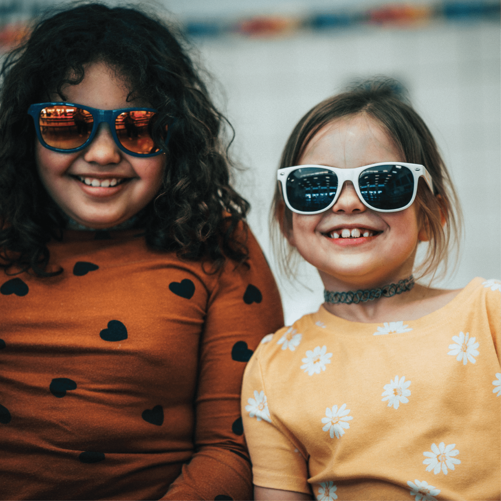 Two girls sitting together wearing sunglasses smile at the camera.