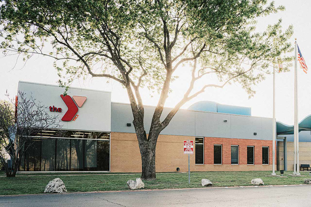 A building with a YMCA logo is seen from a mid-distance. The sun is behind the building, and there are two flagpoles with flags on the right side. A tree is in the foreground.