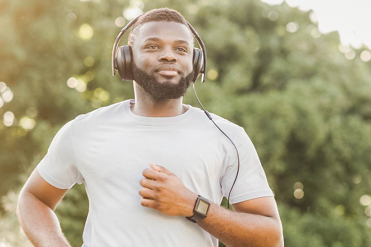 A man in a gray shirt with black headphones on his ears jogs on a trail. Trees are blurred in the background of the photograph